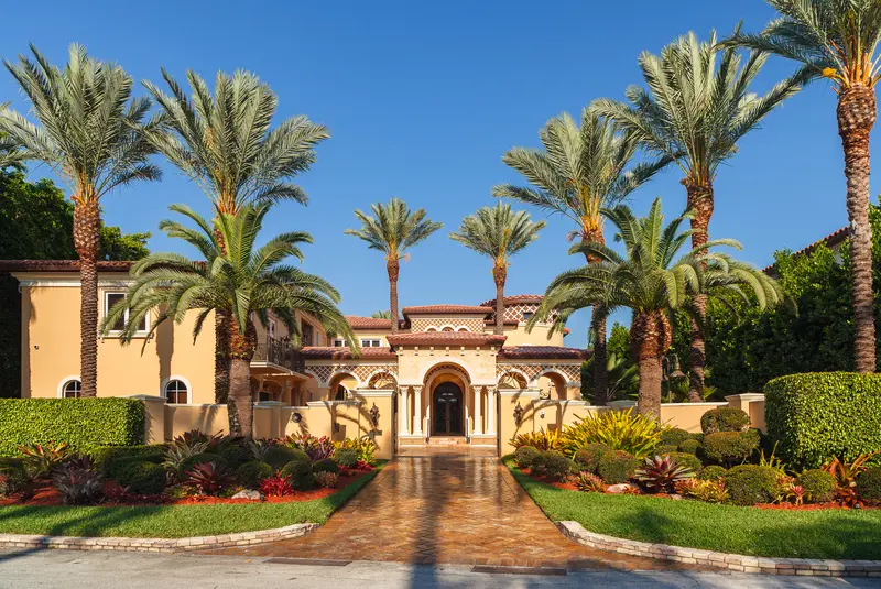 Mediterranean style mansion with detailed landscaping, palm trees, and brick path.