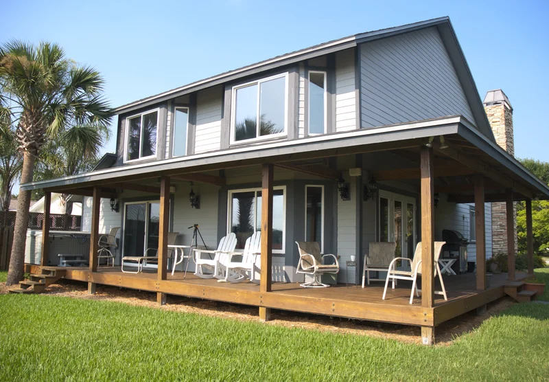 Two-story house featuring a large wooden deck porch surrounded by a green lawn.